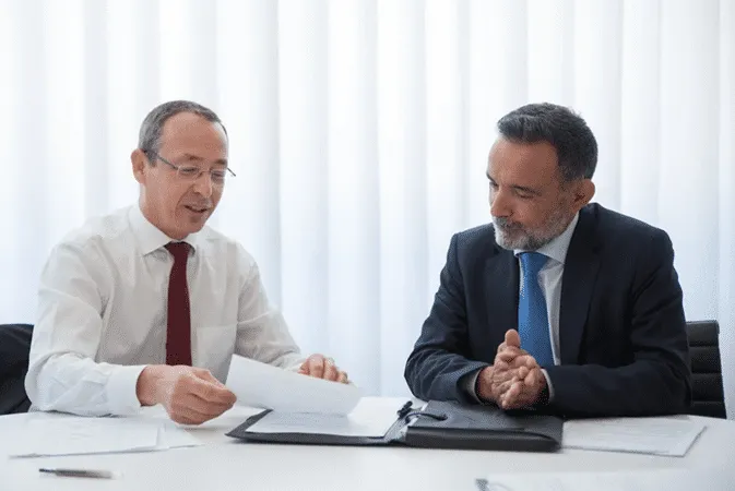 Two men in suits discuss documents at a table, illustrating trust protectors in international estate planning.